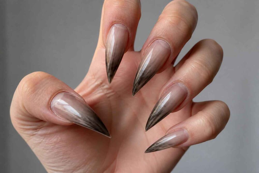 Dłoń z długimi paznokciami stiletto w transparentnym wykończeniu i czarnymi końcówkami na szarym tle